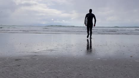 Hombre-Caminando-Hacia-El-Océano-Con-Una-Tabla-De-Surf-Y-Traje-De-Neopreno-Listo-Para-Surfear