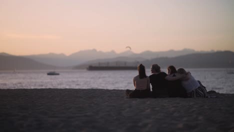 4-friends-sit-on-a-beach-at-sunset,-looking-towards-ocean-and-mountain-landscape