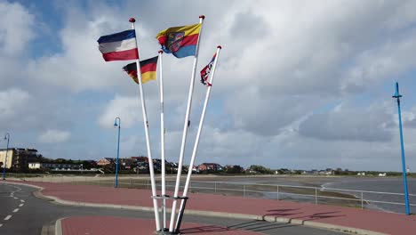 Flagstaffs-with-flags-of-Germany,-Schleswig-Holstein,-Amrum-are-waving-in-heavy-seaside-wind,-clounds-rolling-by-in-the-background,-The-whole-flagstaff-is-visible,-from-top-to-bottom