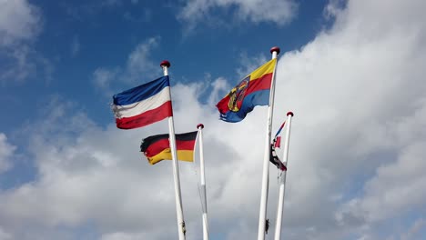 Flagstaffs-with-flags-of-Germany,-Schleswig-Holstein,-Amrum-are-waving-in-heavy-seaside-wind,-clounds-rolling-by-in-the-background,-crop-of-the-top-of-the-flagstaffs