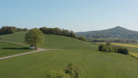 Drone---Aerial-panorama-shot-of-al-lonely-chapel-on-a-field-with-grass-and-a-road-with-an-tractor-and-a-panorama-of-the-seven-mountains---Siebengebirge-30p