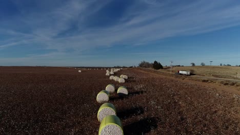 Moving-Drone-Aerial-shot-of-a-Midwestern-cotton-farm-with-fresh-bales-of-harvested-cotton-wrapped-in-bright-yellow-material-against-a-blue-open-sky-Semi-Truck-on-the-road