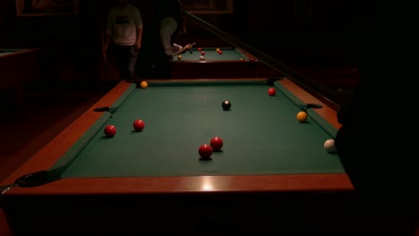Over-the-shoulder,-row-of-two-pool-tables-and-silhouette-of-person-shooting-balls-with-a-cue-stick