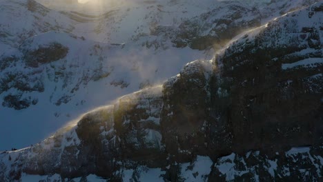 Sunset-drone-approach-of-glistening-snow-blowing-off-of-mountain-ridge