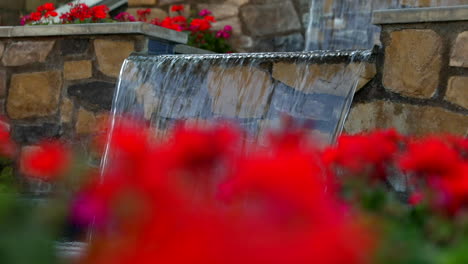 Defokussierte-Ansicht-Von-Roten-Blumen-Mit-Wasserspiel-In-Steinmauer-Im-Hintergrund