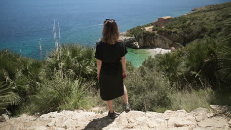 Woman-stands-on-cliff-by-turquoise-blue-beach-in-Sicily,-Italy
