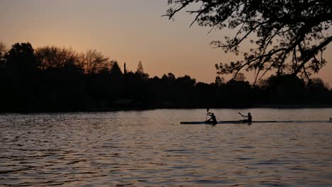 Silhouette-of-two-men-paddling-canoes-in-dam-at-sunset,-golden-hour