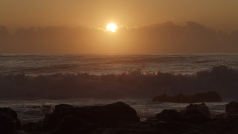 Slow-motion-shot-of-orange,-gold-sun-rising-over-low-cloud-and-breaking-surf