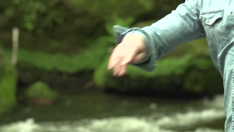 Man-throw-a-stone-pebble-in-the-water-in-nature-in-the-forest-with-water-and-stones-in-the-background-in-slow-motion-close-up-shot
