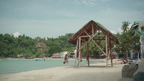 Tourist-Spazieren-Sie-Gerne-Am-Strand-Entlang-In-El-Nido,-Philippinen-Mit-Exotischen-Bäumen-Und-Holzhütten---Perfekt-Für-Sommerferien---Breite-Aufnahme