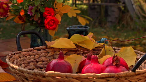 Ripe-red-pomegranates-in-basket-on-garden-table-with-bouquet-of-flowers-in-vase-behind-it