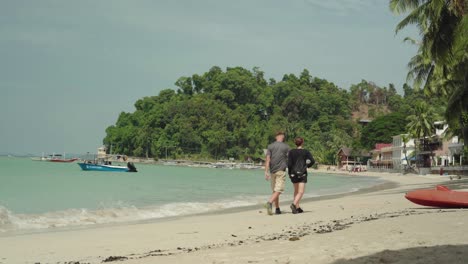 Tourist-Couple-Enjoy-Walking-On-The-White-Sand-Beach-Composed-of-Exotic-Trees-and-Green-Ocean-With-Tourist-Boat-Floating-On-The-Sea---Wide-Shot