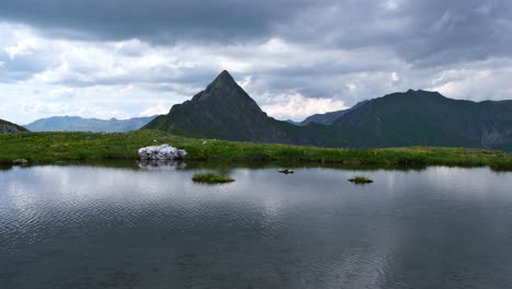 Remote-alpine-lake-with-green-grass-and-pointy-mountain-in-the-background