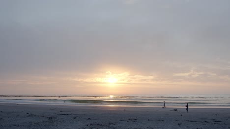 Bewölkter-Sonnenaufgang-Am-Strand,-Während-Die-Menschen-In-Frieden-Mit-Ihren-Hunden-Spazieren-Gehen