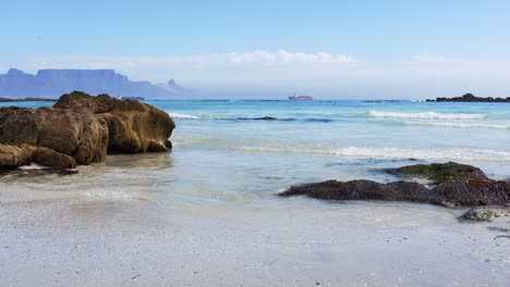 water-swirls-around-the-rock-pool-as-waves-splash-over-rocks,-Table-Mountain-in-the-background-with-a-ship-in-the-distance