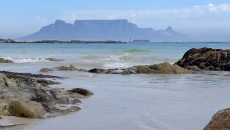 Waves-gently-splash-over-rocks-in-foreground-with-table-mountain-and-a-ship-in-the-distance