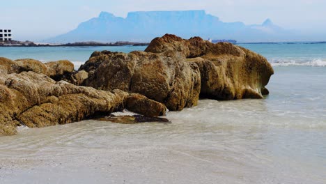 Slow-pan-over-beach-rocks,-Table-Mountain-in-the-background,-waves-lapping-on-the-white-sand