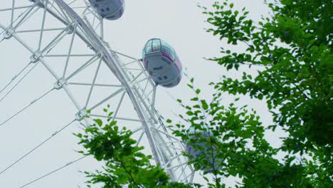 Close-up-crop-shot-of-The-London-Eye-slowly-rotating-on-an-overcast-cloudy-day-with-green-tree-leaves-moving-in-the-breeze