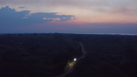 Car-drives-down-narrow-dirt-road-in-Ugandan-wilderness-at-sunset-with-bright-lights
