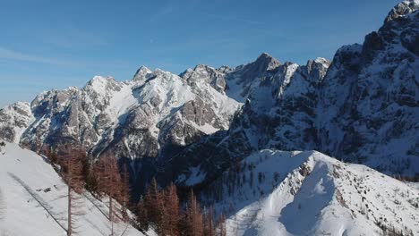 A-snow-covered-rocky-mountain-range-with-pine-trees-beneath-in-Slovenia,-mountain-pass-Vršič---aerial-reveal-shot