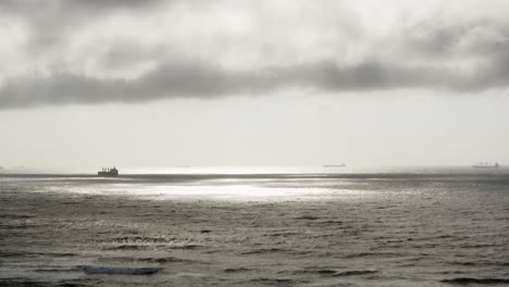 A-calm-ocean-scene-with-dark-clouds-above-and-pools-of-light,-ships-passing-by-slowly-on-the-horizon