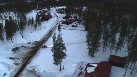 Aerial-flight-to-the-left-over-a-snowy-landscape-of-Fulufjallsbyn-in-Sweden-with-a-snowmobile-turning-of-the-road-at-a-country-road-leading-in-a-forest