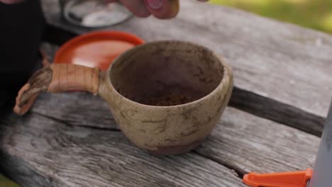 Persons-Hands-Making-Instant-Camping-Coffee-in-a-Wooden-Kup-with-Leather-Handle-on-a-Bench