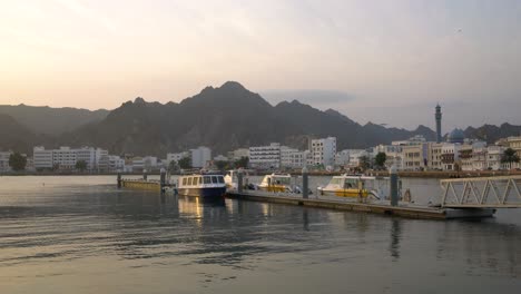 Dock-with-boats-in-Mutrah-corniche-with-birds-flying-and-mountains-in-the-background-during-sunrise