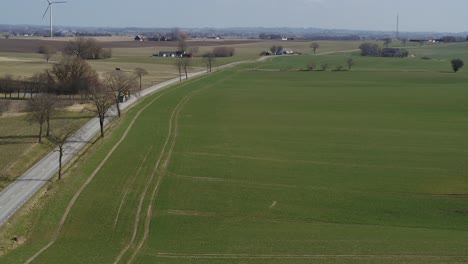 Drohnen-Fliegen-über-Ein-Grünes-Feld-Neben-Einer-Straße-Mit-Einem-Windrad-Im-Hintergrund