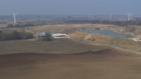 Drohnen-Fliegen-über-Ein-Feld,-Auf-Dem-Ein-Traktor-Pflügt,-Und-Windmühlen-Im-Hintergrund
