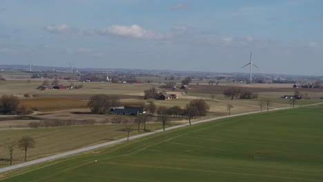 Hohe-Luftaufnahme---Windmühle-Dreht-Sich-Im-Wind-Neben-Einer-Straße-In-Grünen-Feldern-Und-Blauem-Himmel