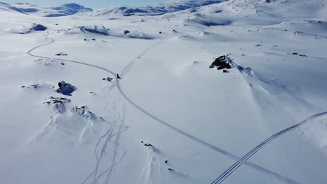 Toma-Aérea-Rastreando-Motos-De-Nieve-A-Través-Del-Paisaje-Montañoso-Cubierto-De-Nieve