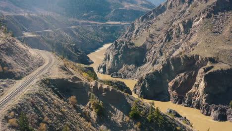 Aerial-rise-up-over-dramatic-valley-river-gorge-with-train-tracks-on-cliffside
