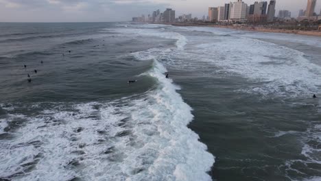 Aerial-view-of-many-surfers,-riding-waves-with-Tel-Aviv-skyline-background---tracking,-drone-shot