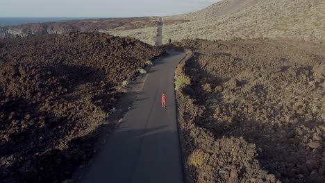 Energetic-woman-in-red-dress-walking-along-a-deserted-road-in-a-valley-between-mountains-and-rocky-shore-on-a-sunny-evening,-Hierro-island,-Canary---drone-view