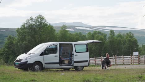 Man-Traveler-And-His-Alaskan-Malamute-Dog-Resting-On-The-Field-Near-Trekanten,-Norway---wide-shot