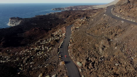 Winding-road-around-a-high-mountain-in-the-Canary-Islands,-which-runs-a-young-woman-in-a-dress,-in-the-background-cliffs-with-the-sea-at-a-relaxing-sunset,-Hierro-island---drone-shot