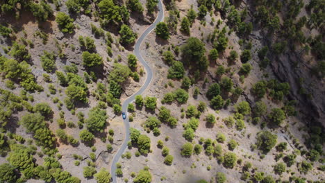 Passenger-car-along-a-winding-road-in-an-arid-landscape-with-green-trees-and-bushes,-Heirro,-Spain---aerial-view-from-a-drone
