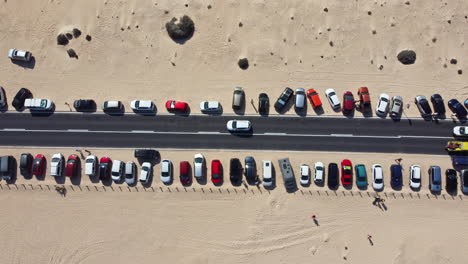 Static-top-down-view-of-parked-cars-by-the-road-with-slow-moving-cars-and-resting-tourists-on-the-yellow-sand-on-a-hot-day
