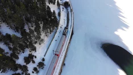 Aerial:-blue-rack-train-in-the-Pyrenees-mountains-travelling-by-a-frozen-lake-and-entering-a-tunnel