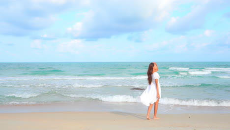 An-attractive-young-woman-in-a-white-sundress-walks-slowly-along-a-sandy-beach-into-the-surf
