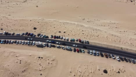 Aerial-view-of-a-road-with-slow-passing-cars-passing-parked-cars-in-a-row,-in-a-sandy-desert-landscape-on-a-sunny-day