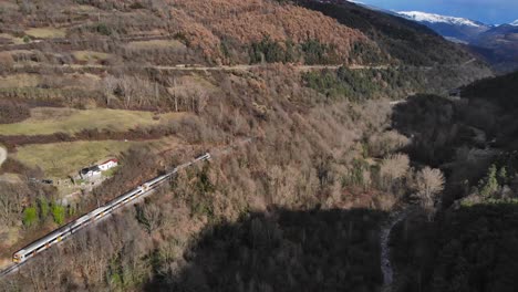 Aerial:-passenger-train-going-through-a-valley-in-a-mountainous-landscape-and-snow-covered-peaks-in-the-background