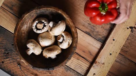 hand-putting-a-red-pepper-next-to-a-wooden-bowl-with-white-mushrooms-inside-on-rustic-wooden-texture-as-background-flat-lay,steady-cam