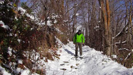 Man-dressed-in-a-green-anorak-walking-along-a-mountain-path-going-among-trees-and-bushes-in-a-snowy-forest,-going-down