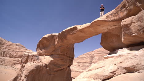 Male-Standing-on-Natural-Arch-in-Desert-Landscape-of-Wadi-Rum,-Jordan-on-Hot-Sunny-Day,-Panorama