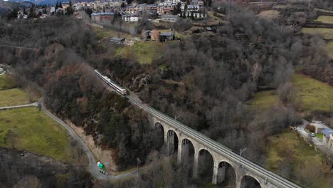 Aerial:-passenger-train-crossing-a-stone-bridge-in-a-mountainous-landscape,-mountainous-town-and-snow-covered-peaks-in-the-background