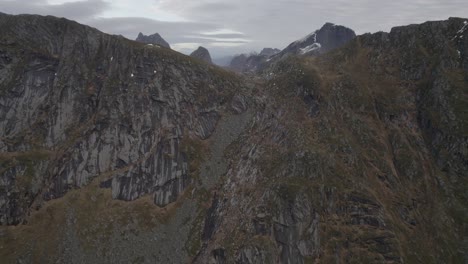 Aerial-view-of-a-rising-up-a-mountain-wall,-revealing-alpine-peaks,-in-cloudy-Lofoten,-Norway---ascending,-drone-shot