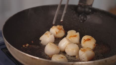 Scallops-cooking-and-sizzling-on-frying-pan-with-oil-popping-and-splattering,-chef-flipping-sides-with-a-pair-of-tongs,-tossing-and-swirling-the-pan,-culinary-scene-filled-with-aromatic-smokes