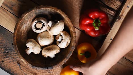 hand-putting-two-yellow-pepper-next-to-a-red-pepper-and-wooden-bowl-with-white-mushrooms-inside-on-rustic-wooden-texture-as-background-flat-lay,-steady-cam